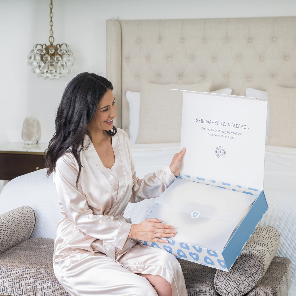 Woman in a satin robe holding The Longevity Pillow product box in a bedroom setting.
